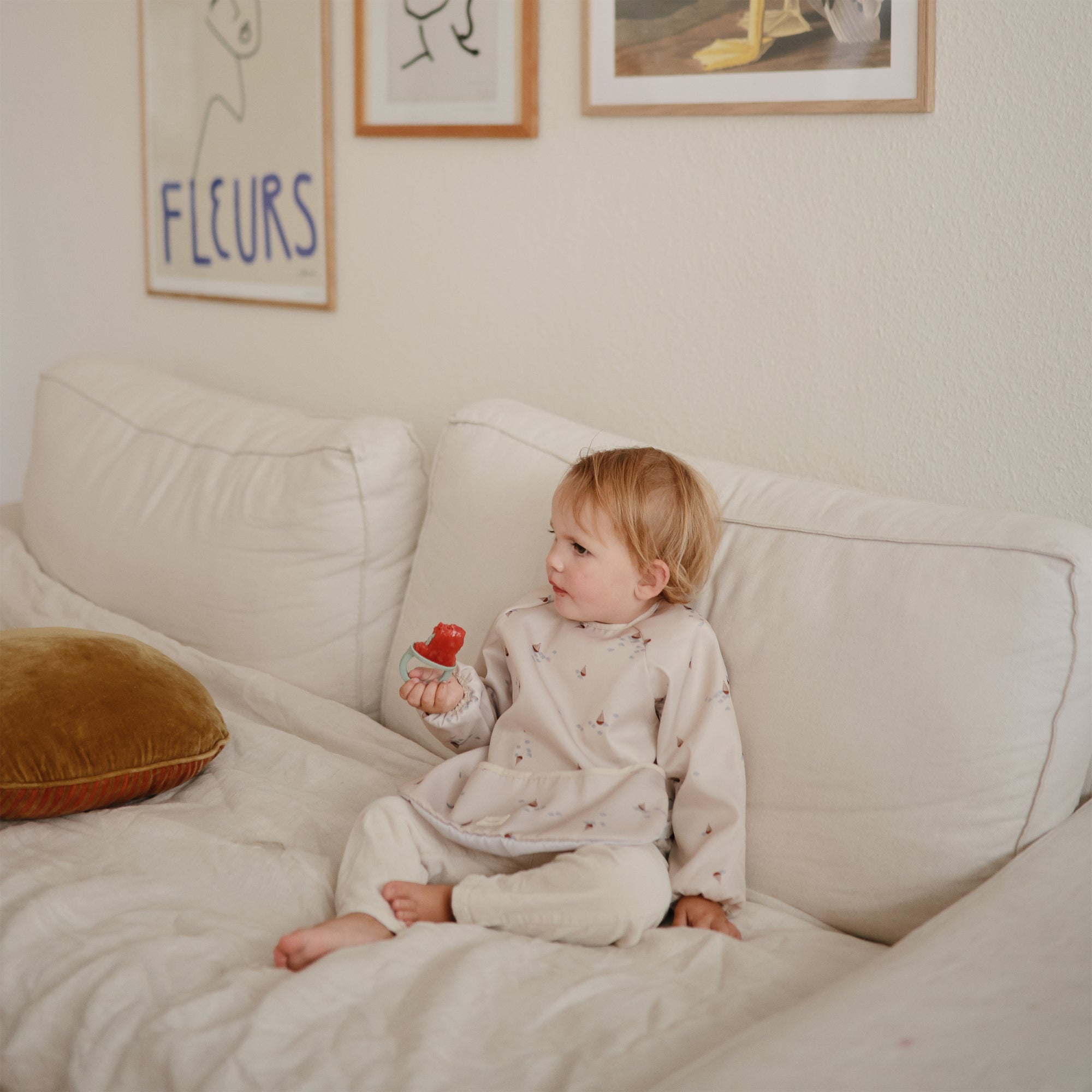 Toddler in a mushie Long Sleeve Bib eats a strawberry on a white couch with art behind.