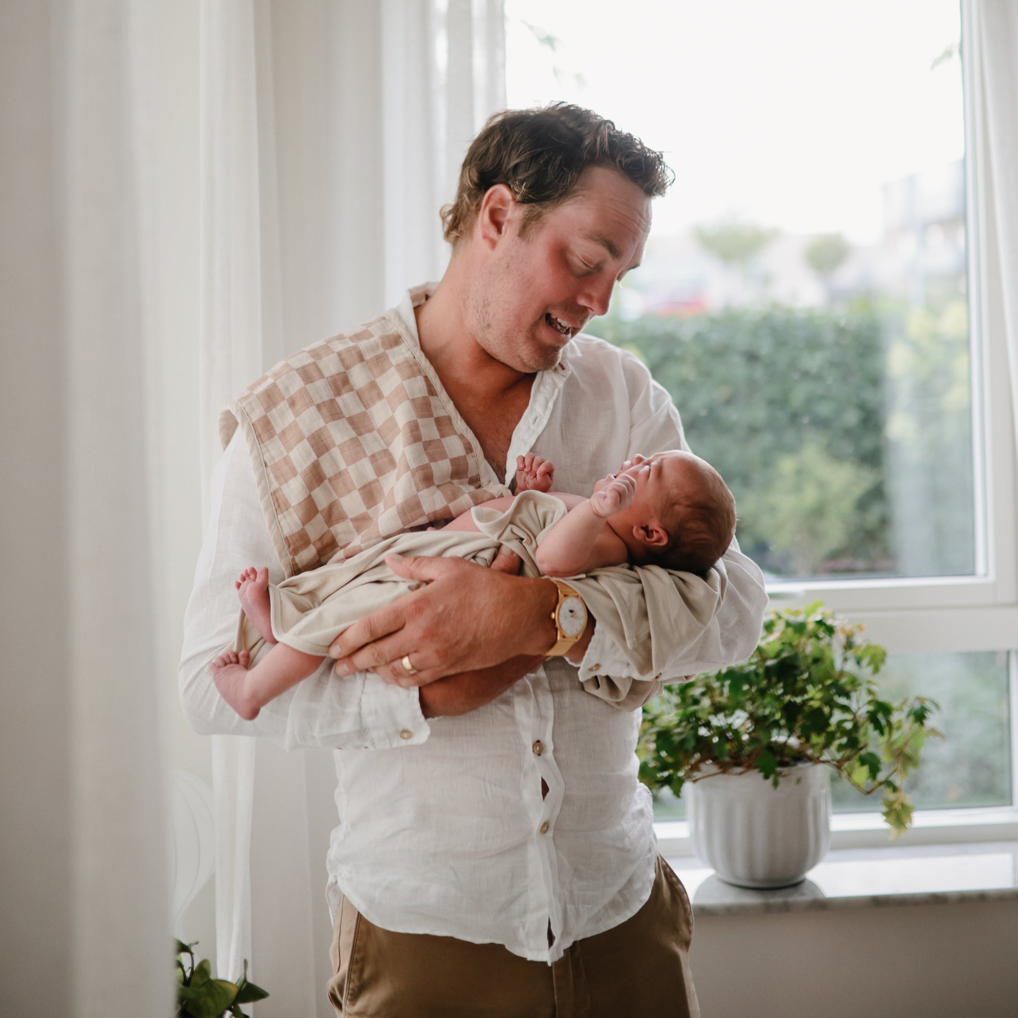 A man in a white shirt smiles as he gently holds a newborn wrapped in the mushie Stretchy Swaddle near a window with green plants on the sill, both bathed in soft natural light.