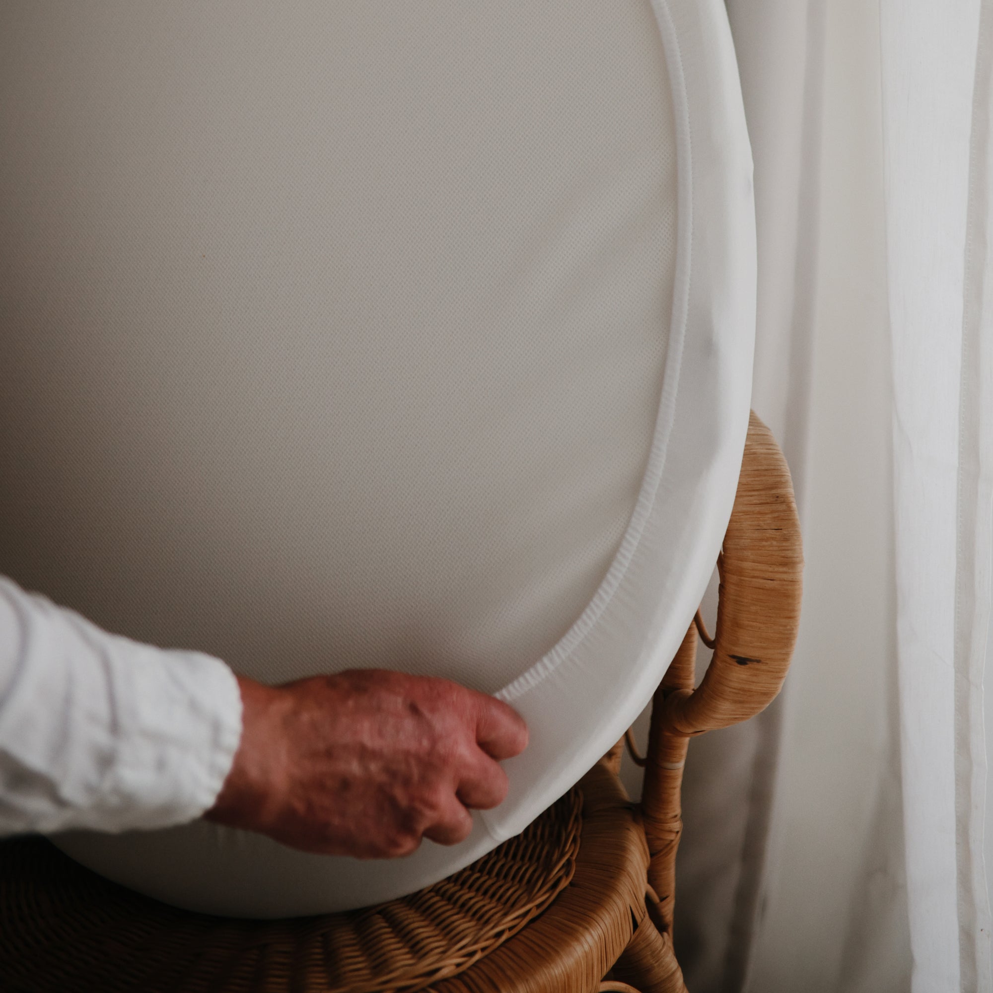 A hand places the mushie Stretchy MINI Crib Sheet, made from breathable organic cotton, onto a woven rattan chair beside a sheer white curtain, creating a cozy, inviting space.