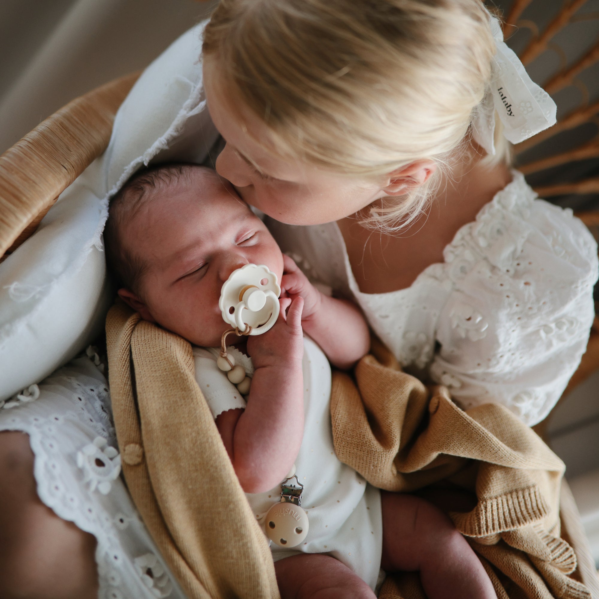 A young girl kisses a baby swaddled in tan with a FRIGG Daisy Natural Rubber Pacifier.