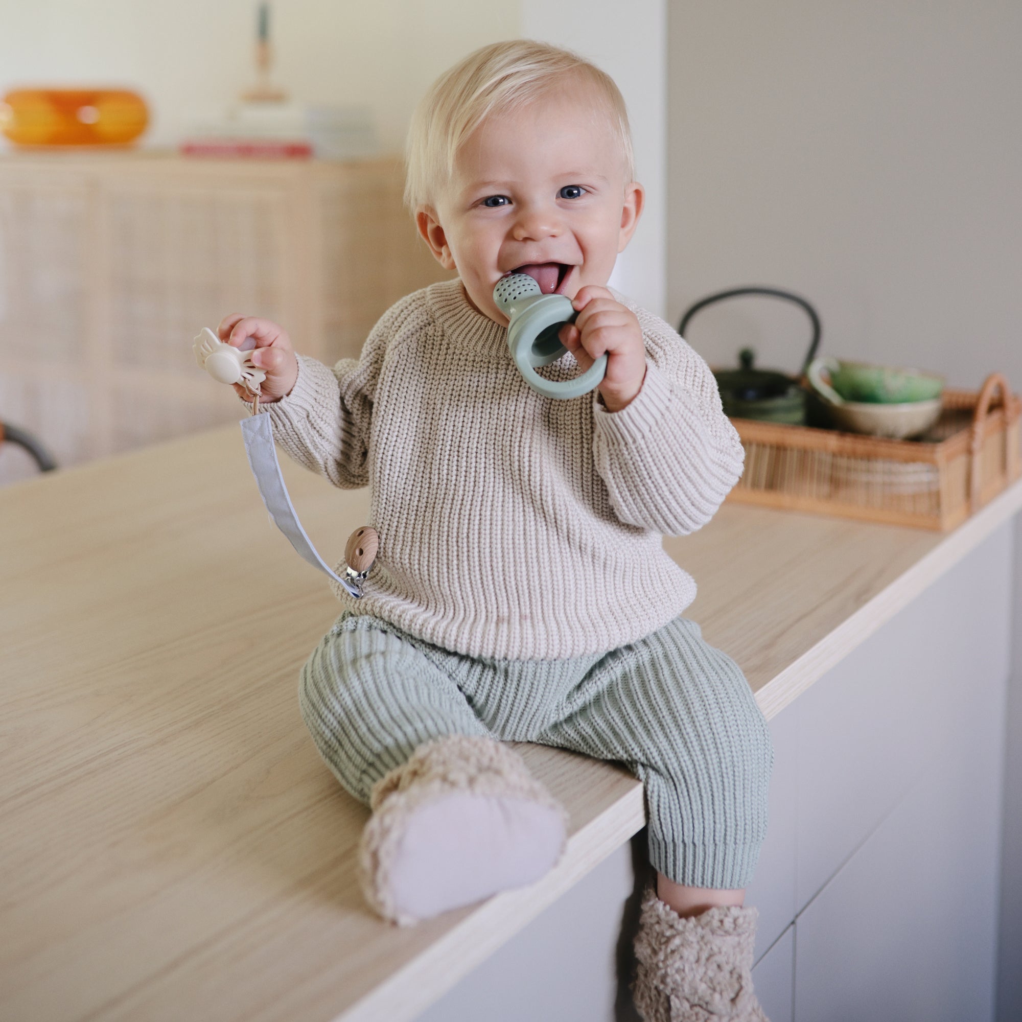 A smiling blond toddler sits on a kitchen counter in a cream sweater and green pants, holding the FRIGG Butterfly Anatomical Silicone Pacifier 2-Pack (0-6 Months) for comfort.