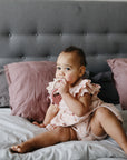 Baby in a pink dress chews the mushie Fairy Teething Ring on the bed.