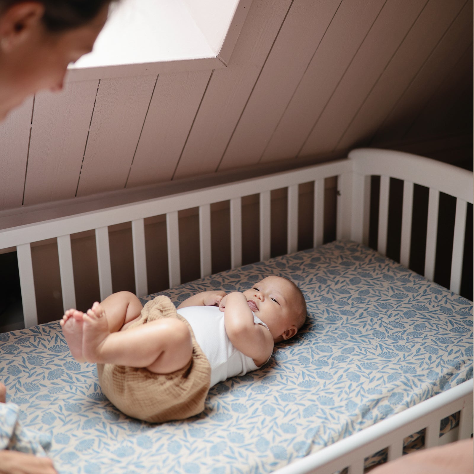A baby on a mushie Extra Soft Muslin Crib Sheet looks up at an adult from the crib.