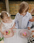 Two kids sit at a table with cakes on mushie Round Dinnerware Plates with pink flowers.