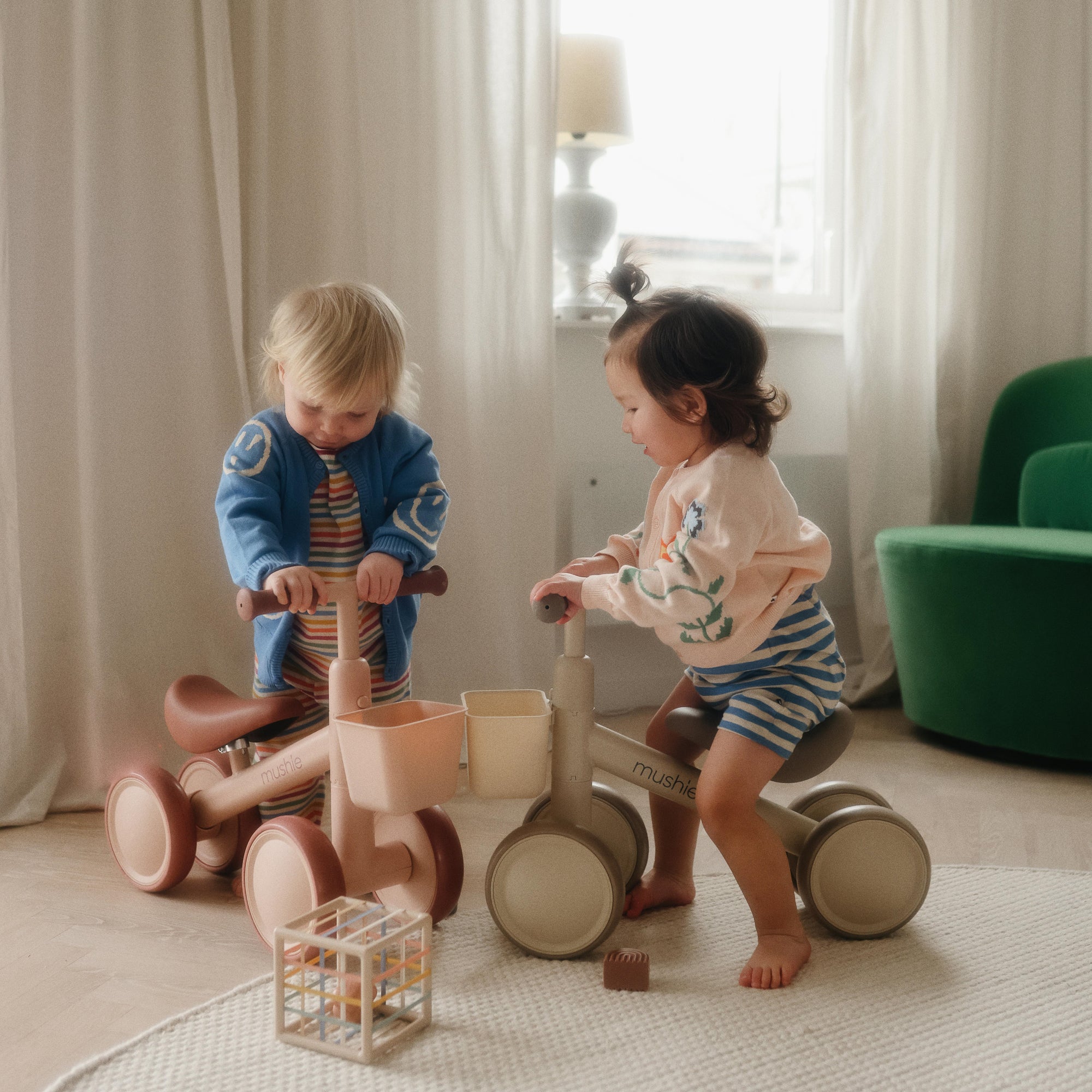 Two young children ride a mushie Mini Toddler Bike indoors near a green chair.