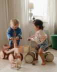 Two young children ride a mushie Mini Toddler Bike indoors near a green chair.