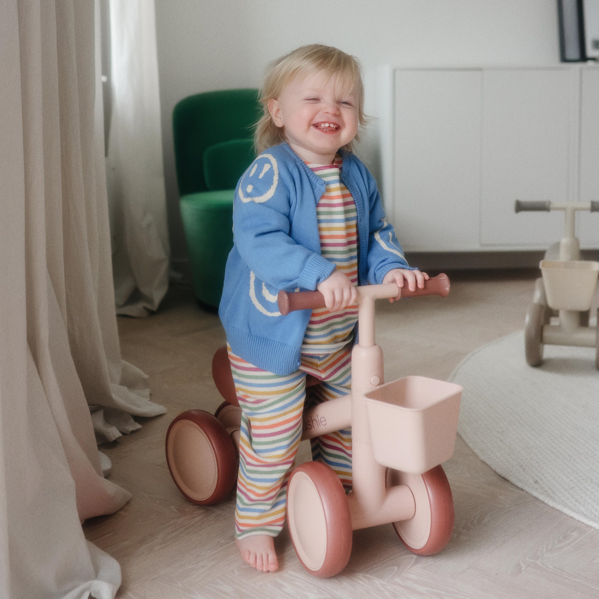 Smiling toddler rides a mushie Mini Toddler Bike indoors, wearing colorful clothes.