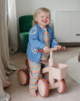 Smiling toddler rides a mushie Mini Toddler Bike indoors, wearing colorful clothes.