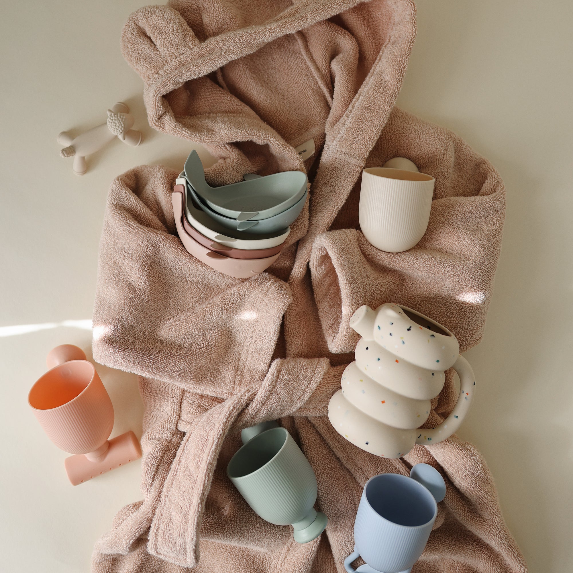 Pink bathrobe with Mushie Whale Bath Stackers, pastel cups, and bowls arranged on top.