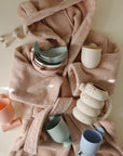 Pink bathrobe with Mushie Whale Bath Stackers, pastel cups, and bowls arranged on top.