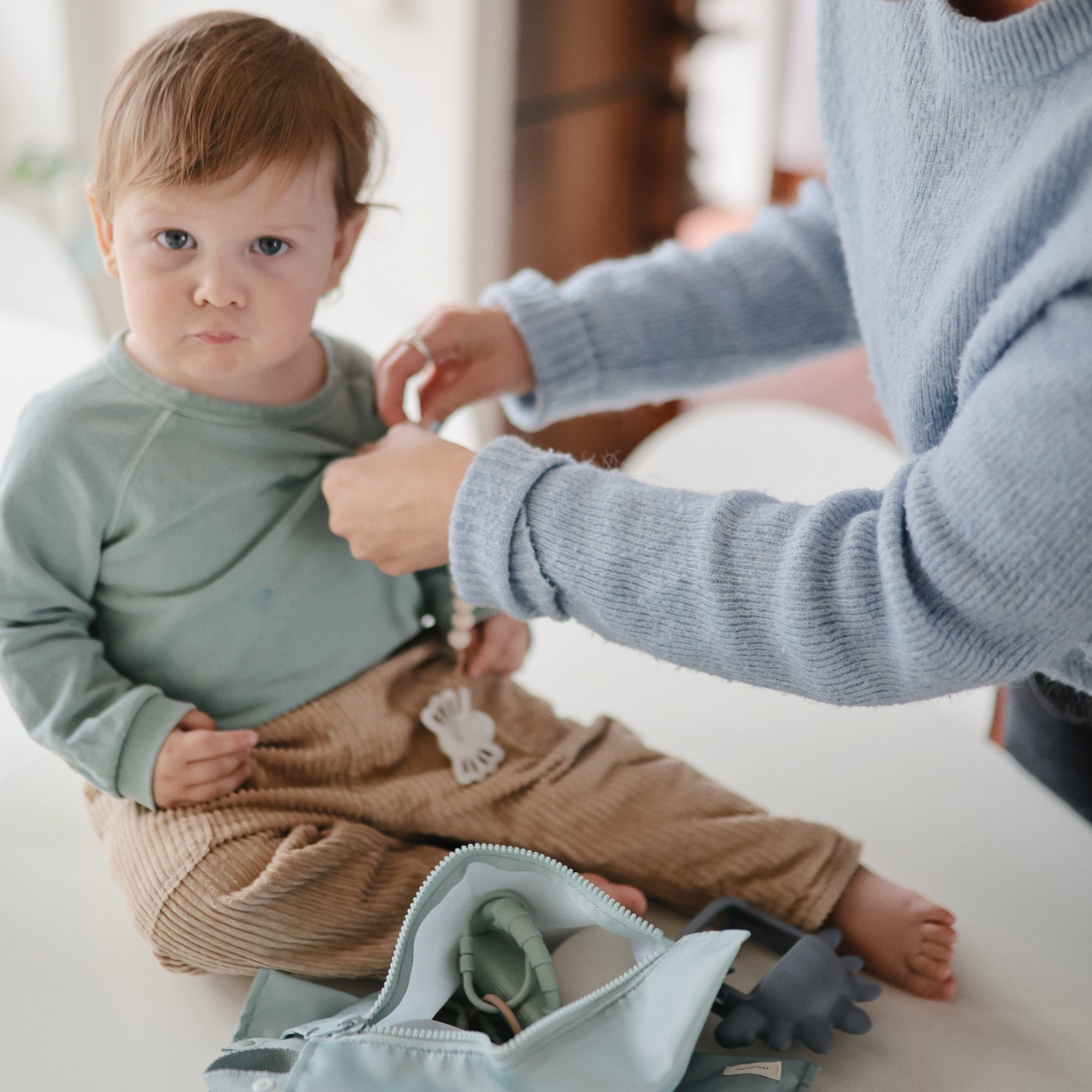A young child sits on a white surface as an adult helps with the child's mint green shirt. Nearby is a light blue bag containing the FRIGG Lucky Symmetrical Silicone Pacifier 2-Pack (0-6 Months) from FRIGG for baby oral development.