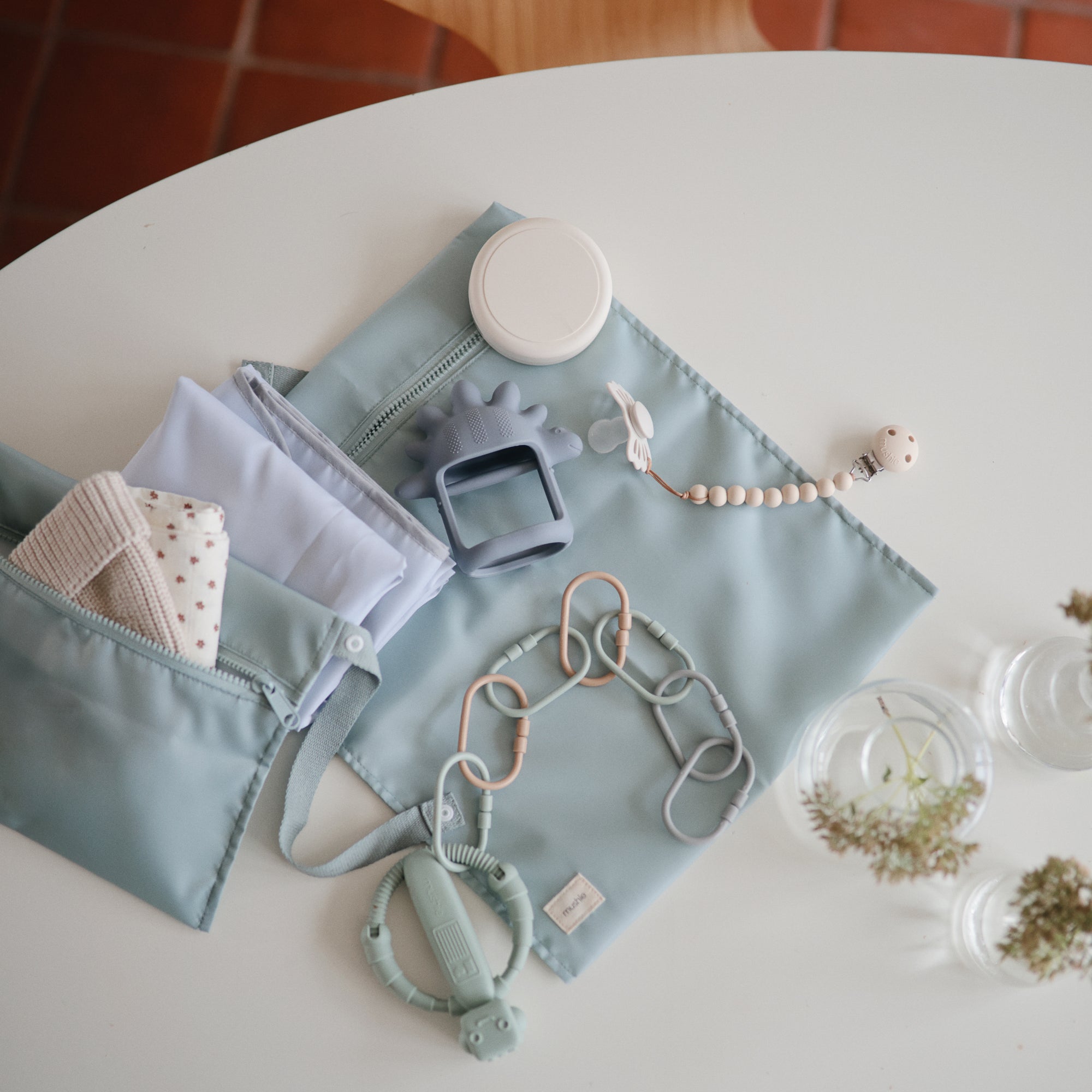 A light blue diaper bag on a white round table holds a folded cloth, pacifier clip, teether, small container, and silicone toys. Nearby are two glass cups with small flowers and a FRIGG Baby’s First Pacifier Moonlight Sailing (Cream) 4-Pack.