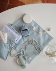 A light blue diaper bag on a white round table holds a folded cloth, pacifier clip, teether, small container, and silicone toys. Nearby are two glass cups with small flowers and a FRIGG Baby’s First Pacifier Moonlight Sailing (Cream) 4-Pack.