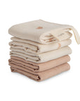A neatly stacked set of six mushie Muslin Cotton Washcloths in assorted beige and brown tones, featuring a hanging loop and rainbow embroidery on the top cloth.