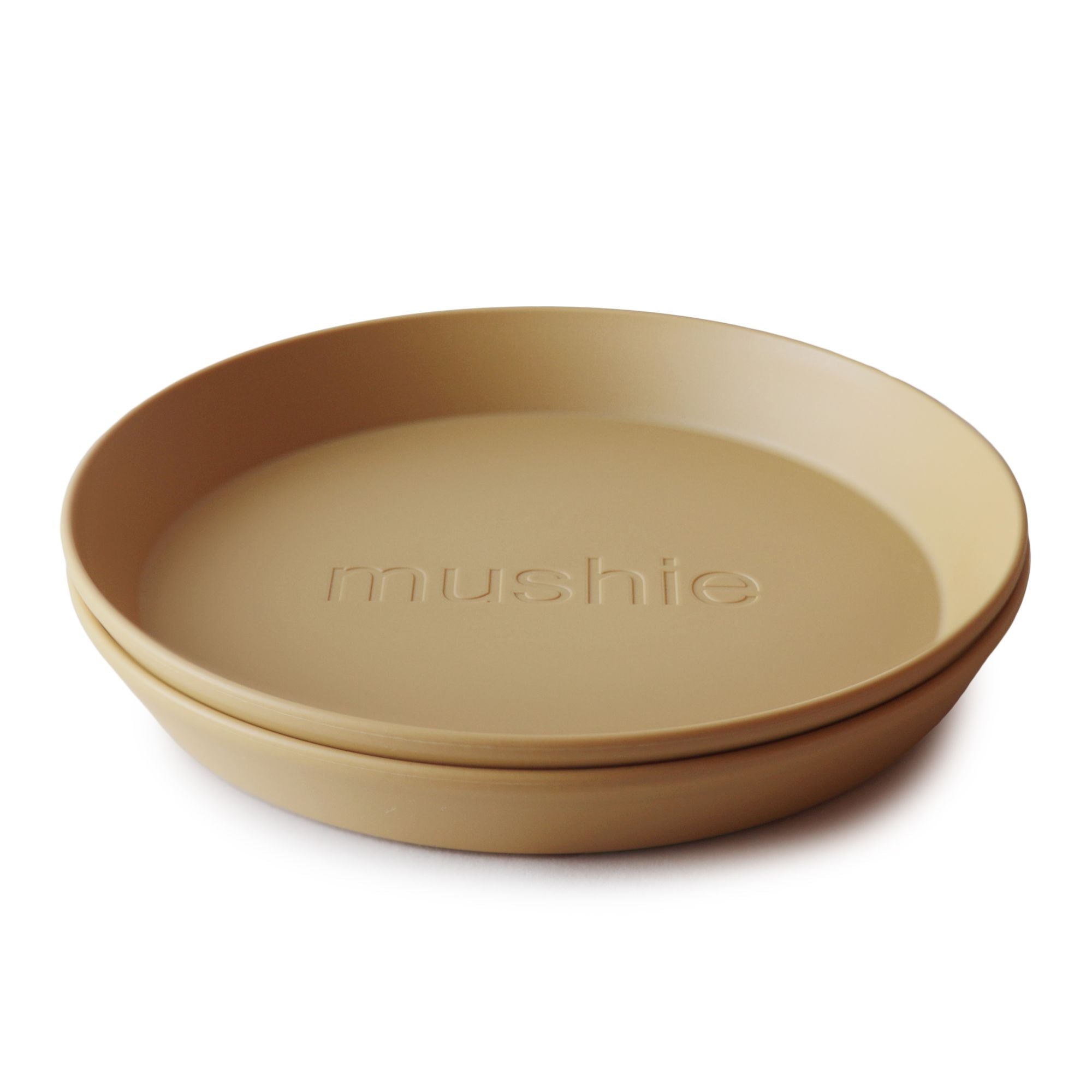 Two stacked tan Round Dinnerware Plates by mushie, made from non-toxic plastic with "mushie" embossed in the center, displayed against a white background.