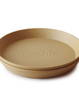 Two stacked tan Round Dinnerware Plates by mushie, made from non-toxic plastic with "mushie" embossed in the center, displayed against a white background.