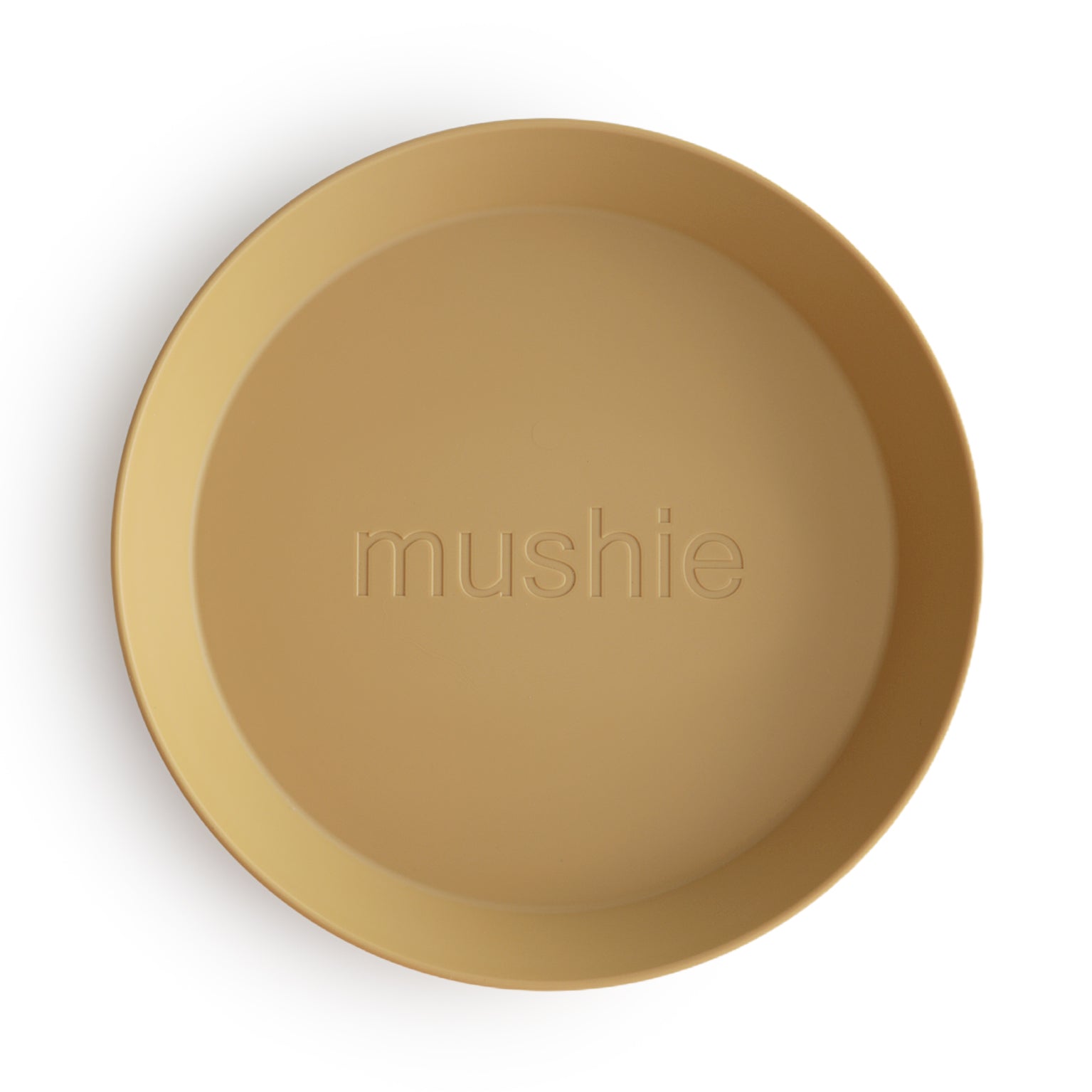 A round, tan mushie Round Dinnerware Plate made of non-toxic plastic features the “mushie” logo in the center and is photographed on a white background—ideal for everyday use.