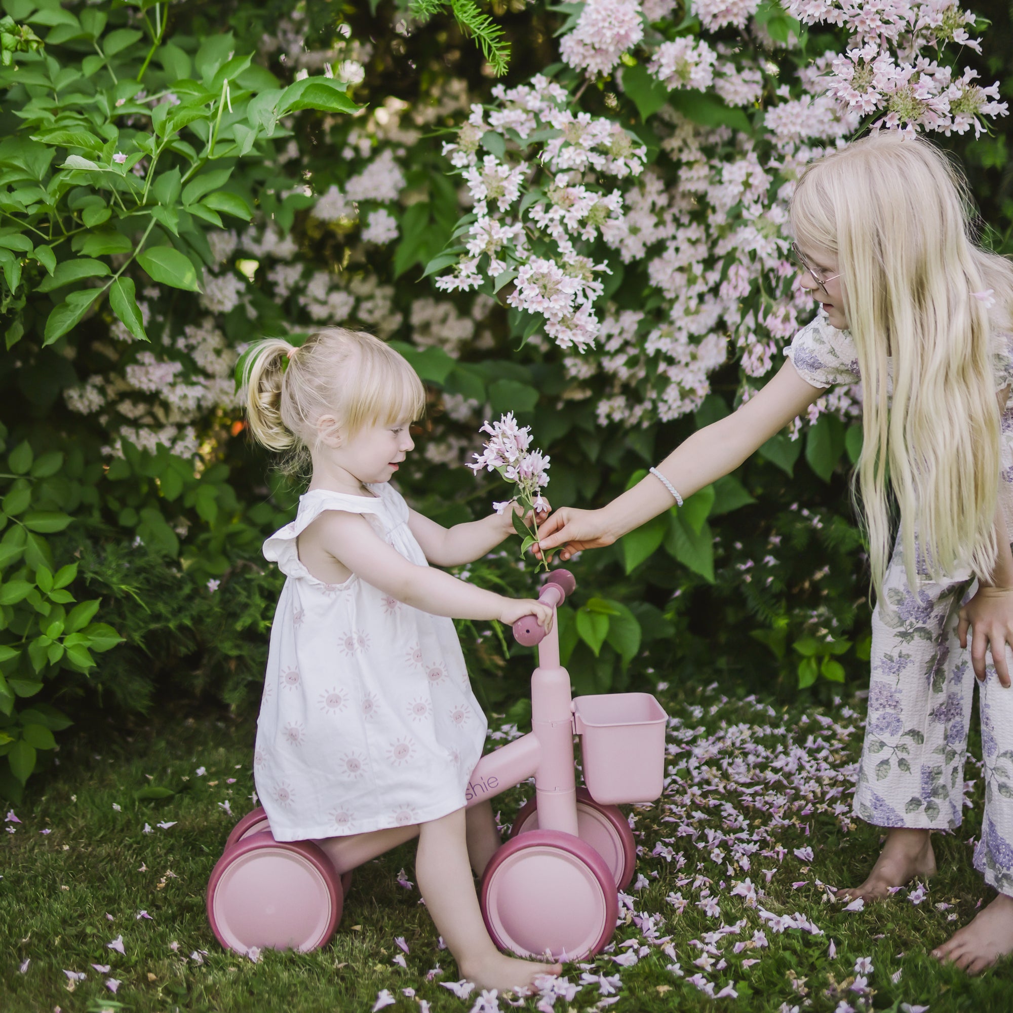 Two blonde girls share flowers by a mushie Mini Toddler Bike in a lush garden.