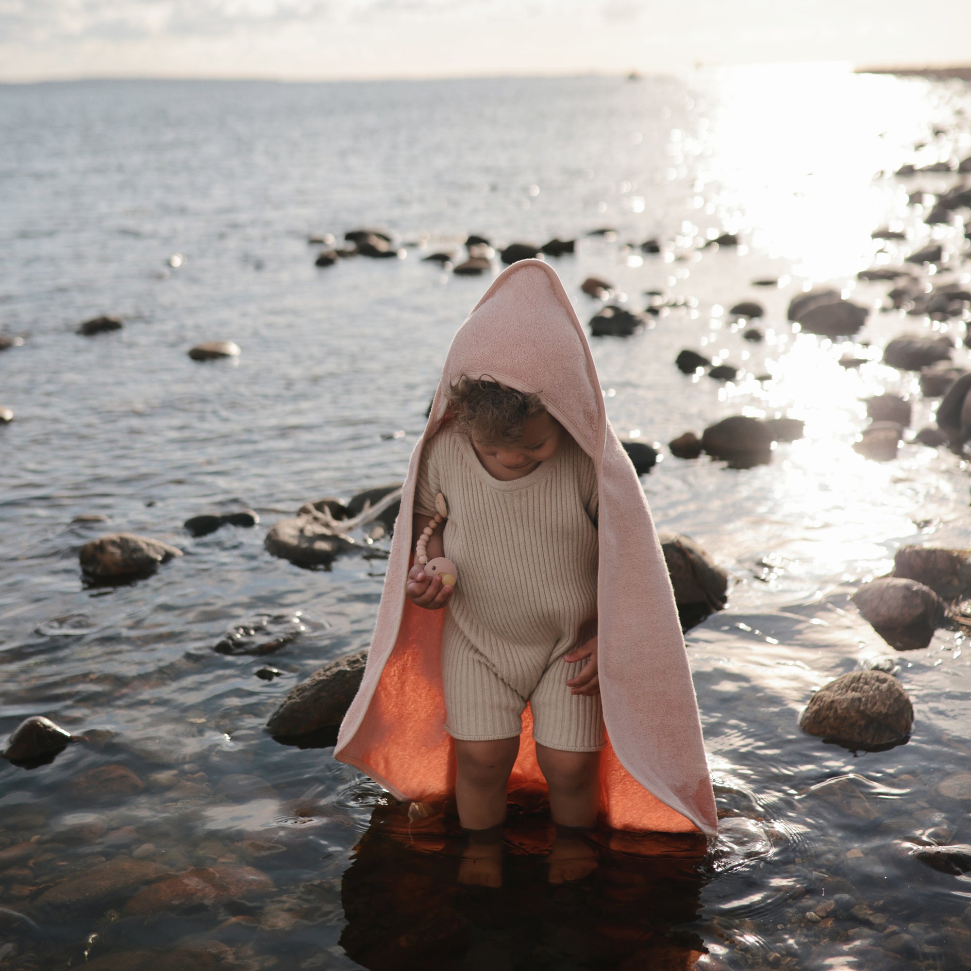 A young child in a beige outfit and pink hooded towel stands among rocky shallows, holding a Mushie FRIGG Rope Natural Rubber Pacifier as sunlight reflects off the water and the shimmering sea glows in the background.