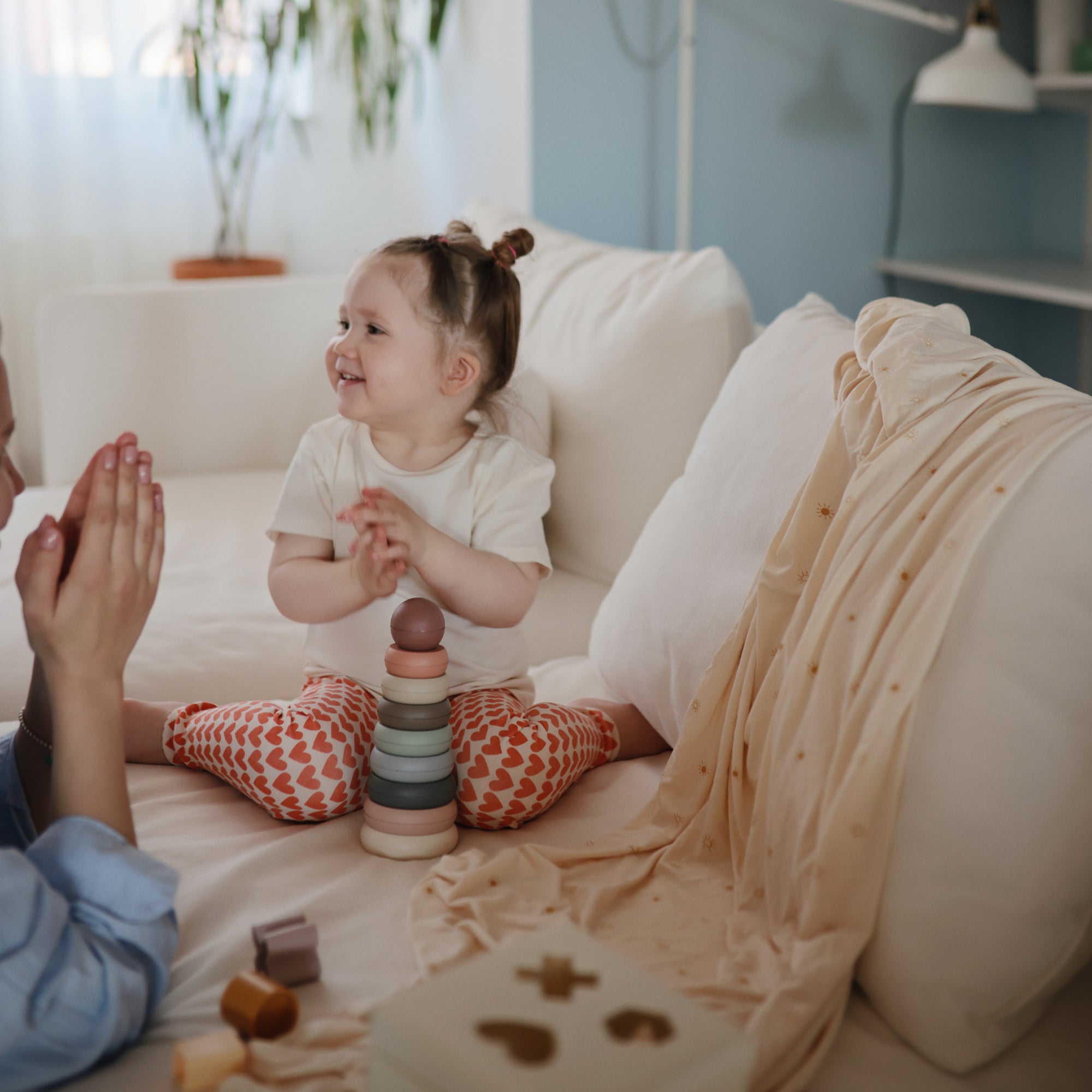 A young child sits on a white couch, smiling and clapping hands with an adult. Colorful stacking toys are in front, and a Mushie Ribbed Baby Blanket is draped nearby in the softly lit, cozy room.