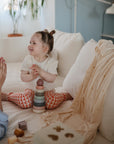 A young child sits on a white couch, smiling and clapping hands with an adult. Colorful stacking toys are in front, and a Mushie Ribbed Baby Blanket is draped nearby in the softly lit, cozy room.