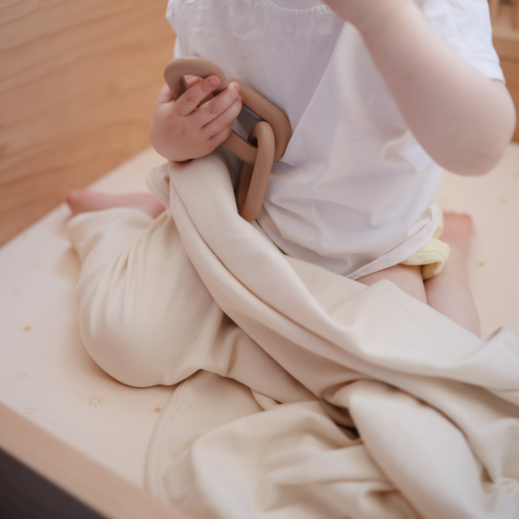 A small child sits on a bed holding the Mushie Ribbed Baby Blanket in beige and a ring-shaped teething toy, dressed in a white shirt and light shorts.