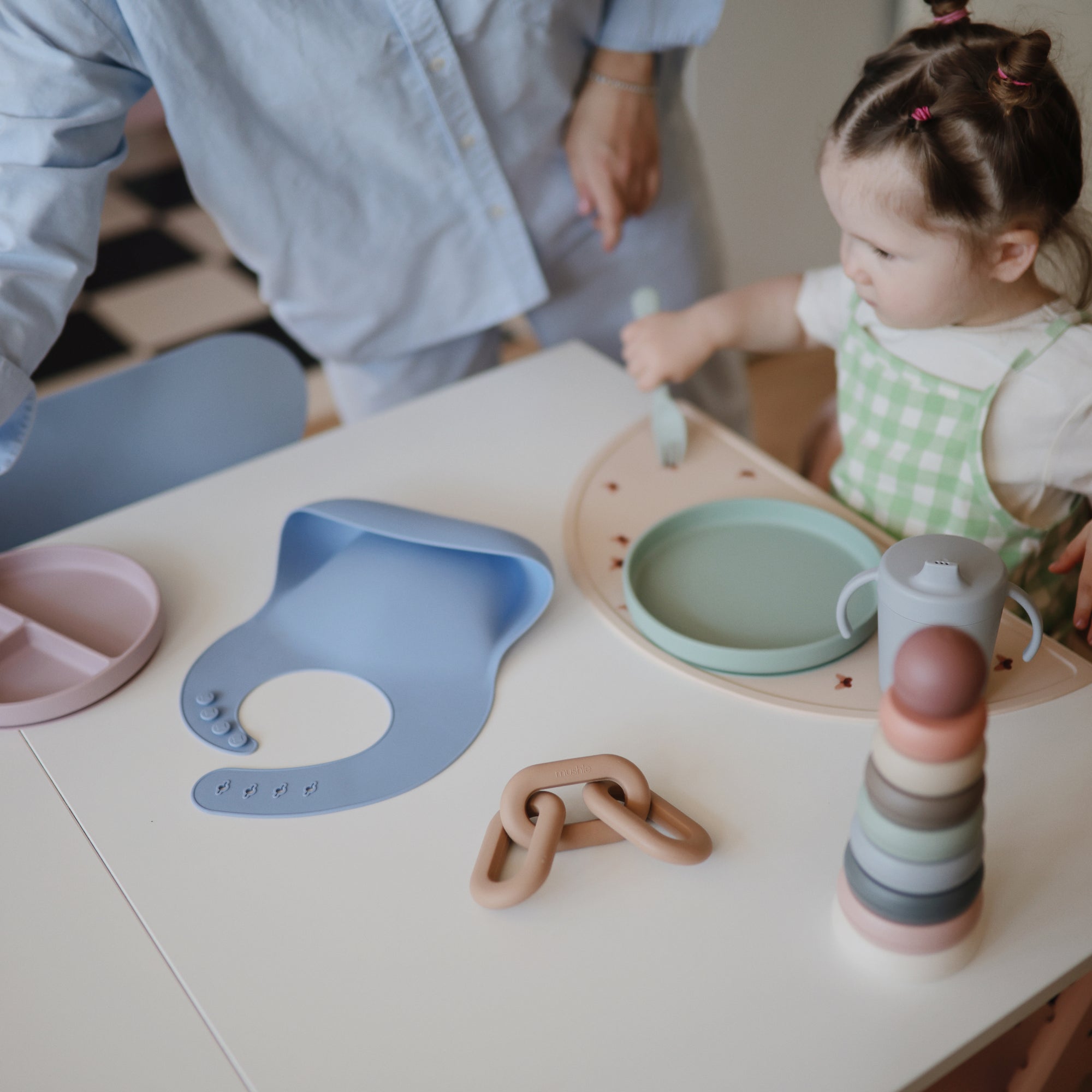 Toddler at table with mushie pastel dishes, bib, cup, and Stacking Rings Toy; adult nearby.