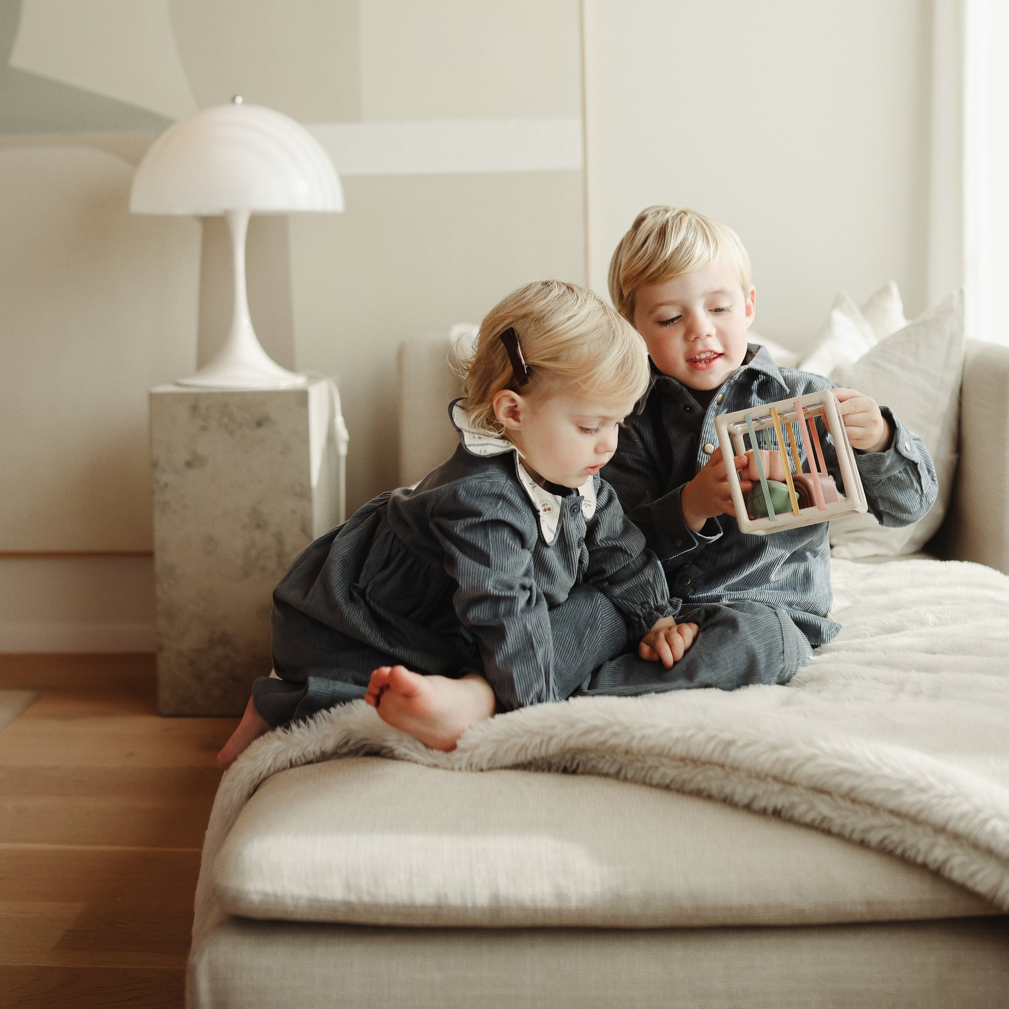 Two kids play on a couch with the mushie Elastic Shape Sorter, building fine motor skills.