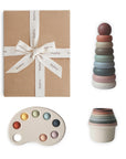 The mushie Playtime Gift Set ($48 value) comes neatly wrapped with a white ribbon and includes pastel sensory toys—a stacking toy, nesting cups, and a paint palette-shaped toy—to help develop fine motor skills. Shown on a white background.
