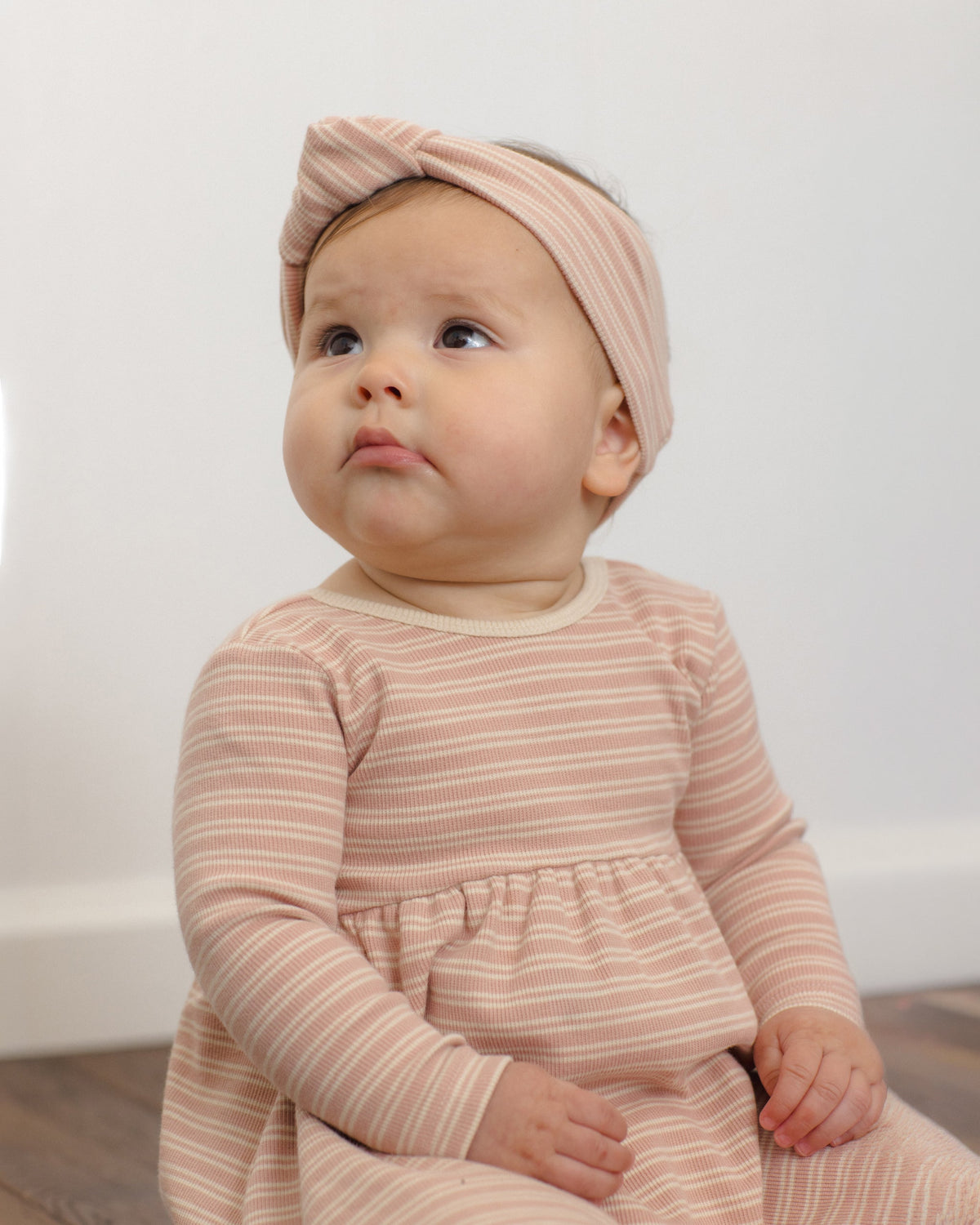 Baby in Rylee + Cru Ribbed Long Sleeve Dress Dusty Rose Microstripe sits on floor looking up.