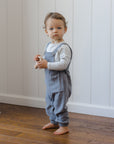 Toddler in gray overalls and Rylee + Cru Long Sleeve Tee Cloud Stripe stands barefoot with toy.