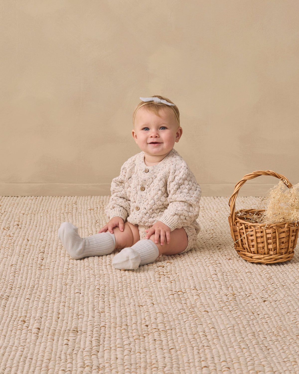 Baby in Rylee + Cru Bubble Knit Cardigan sits on a rug by a straw-filled wicker basket.