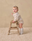 Happy baby in Rylee + Cru Bubble Knit Cardigan holds stool, stands on a textured rug.