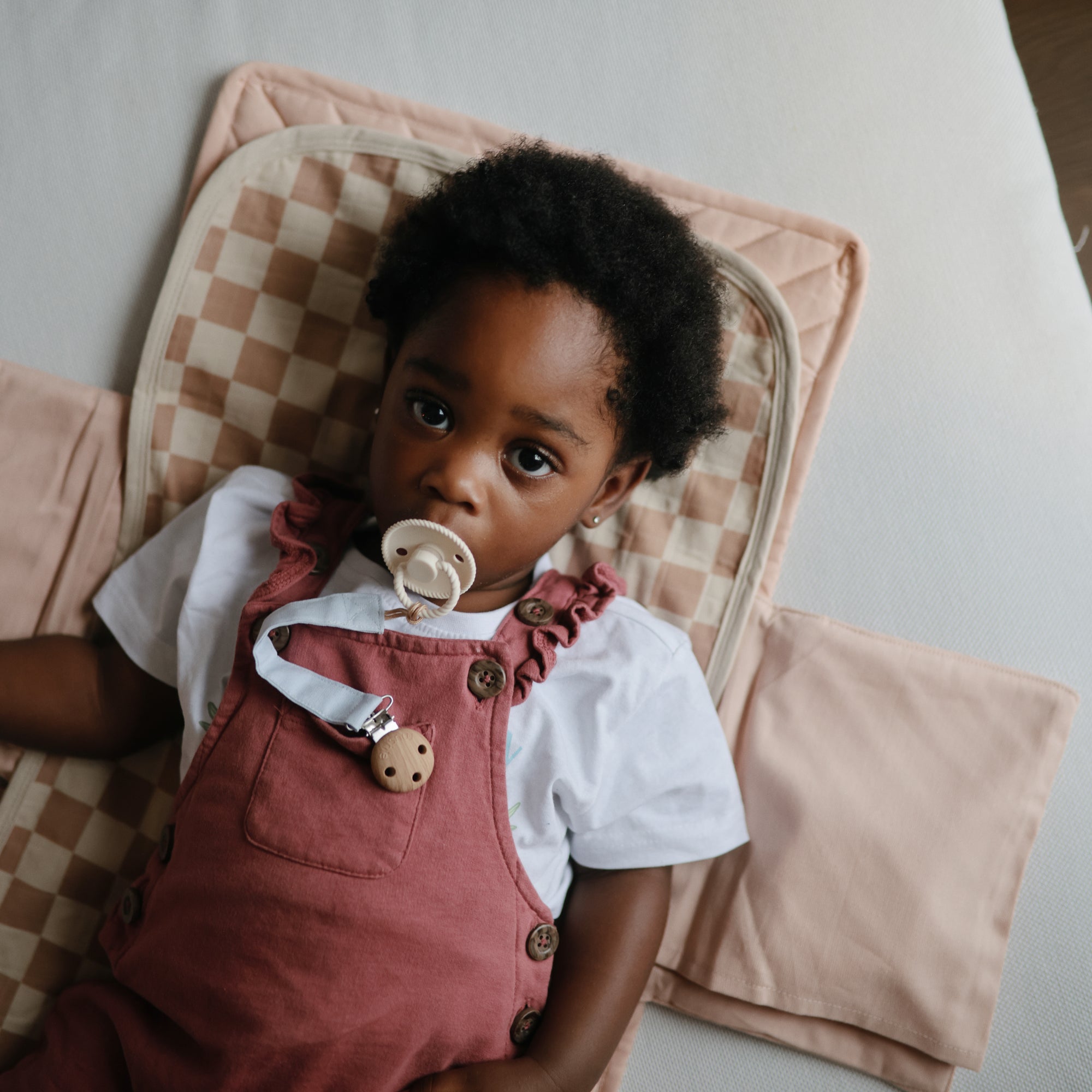 A young child with curly hair lies on a pink and beige checkered mat, wearing a white shirt and pink overalls, looking up with a pacifier held securely by the mushie Linen Pacifier Clip 2-Pack.
