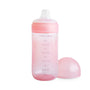 The mushie Soft Spout Sippy Cup in pink features a clear nipple, measurement markings, and comes with a matching cap beside it—ideal for toddlers moving from bottles. Set against a white background.