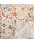 The mushie Organic Cotton Muslin Swaddle Blanket in Space features a colorful space-themed pattern with planets, rockets, stars, and moons on a white background. One corner is folded up to reveal the reverse side.