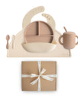 The mushie Stage 2 Natural/Shifting Sand Feeding Kit, featuring a beige silicone plate, bowl, bib, training cup with straw, spoon, and fork on a mat, is shown above a brown gift box with a white ribbon.