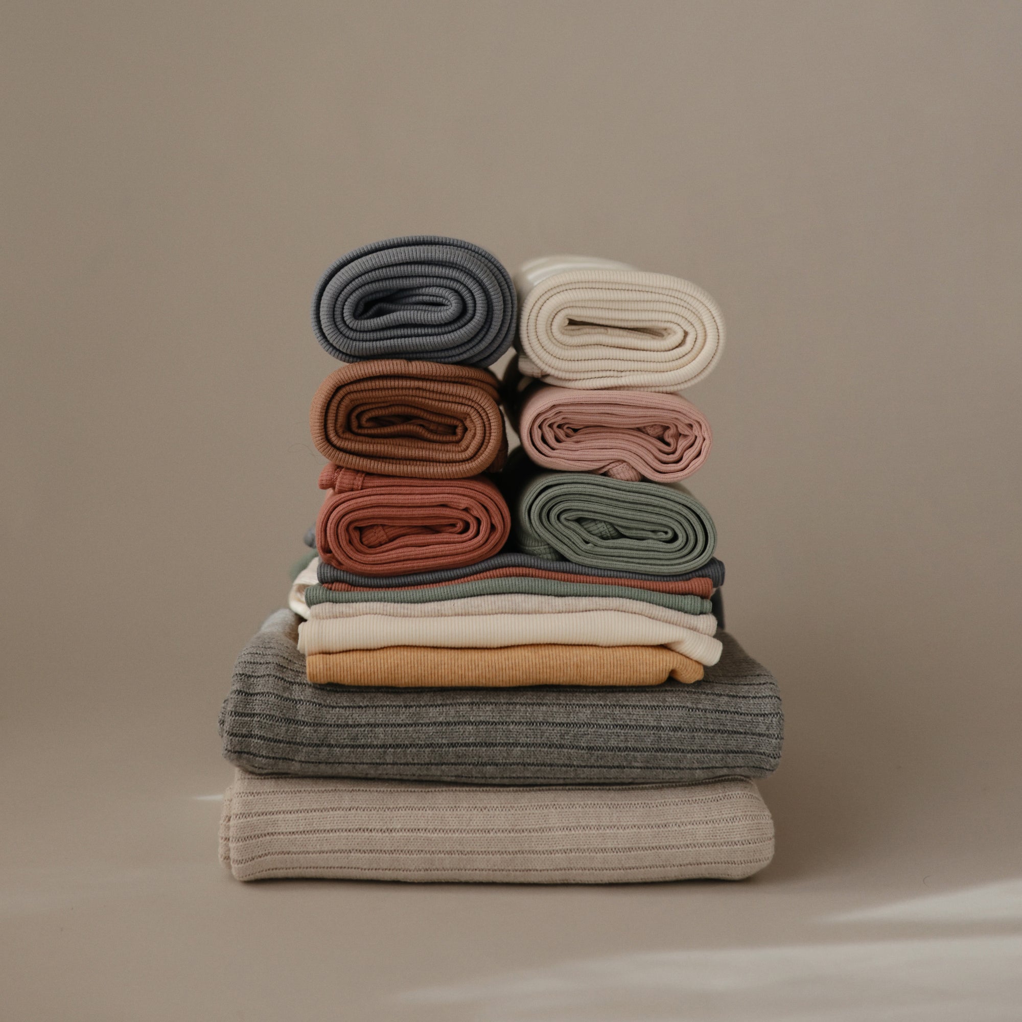 A stack of Mushie Ribbed Baby Blankets in grey, beige, cream, pink, and sage offers comfort and breathability; the soft blankets are folded and rolled, displayed against a taupe background.