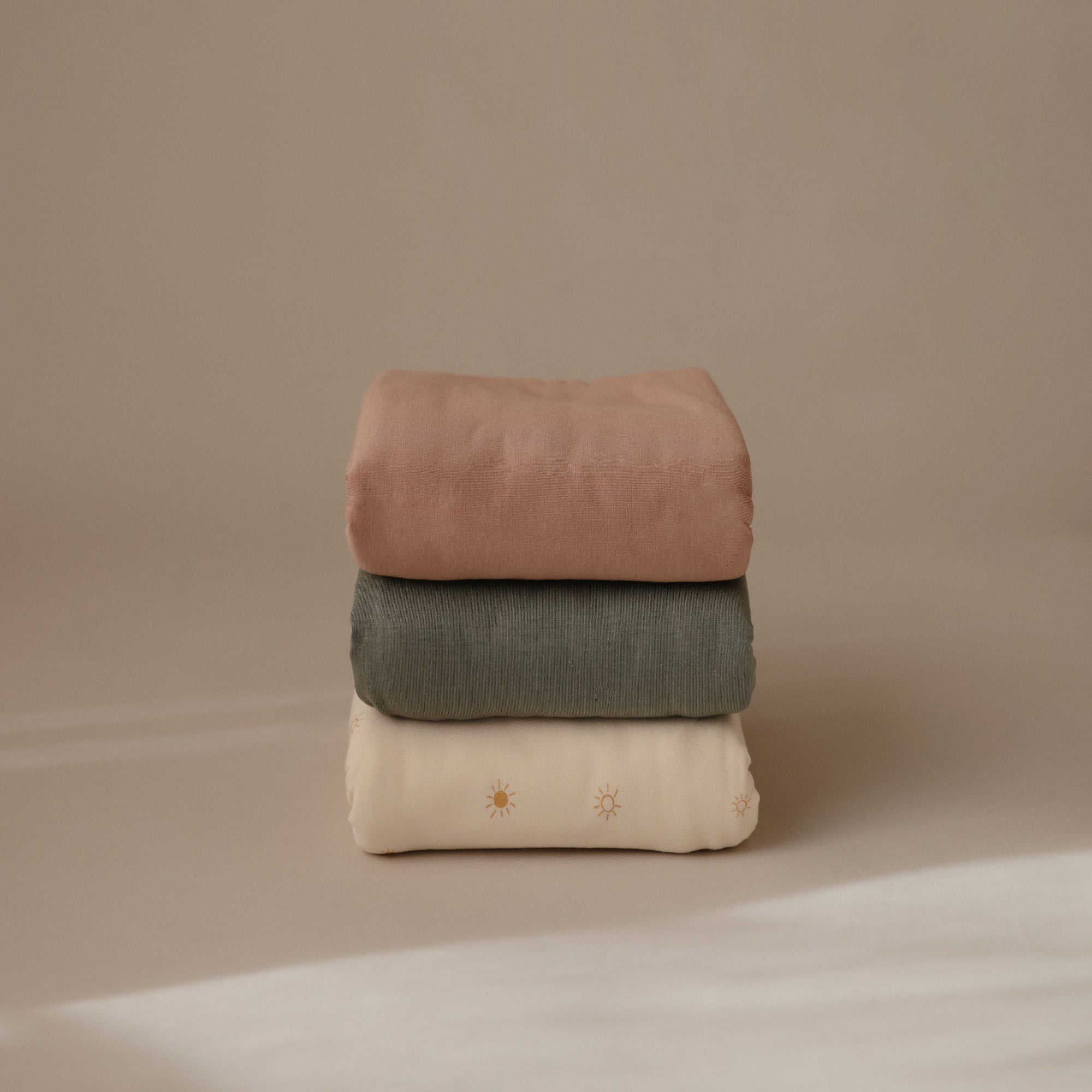 A stack of three blankets against a beige background: mushie Stretchy Swaddle in light brown on top, olive green blanket in the middle, and a cream baby blanket with small sun patterns on the bottom.