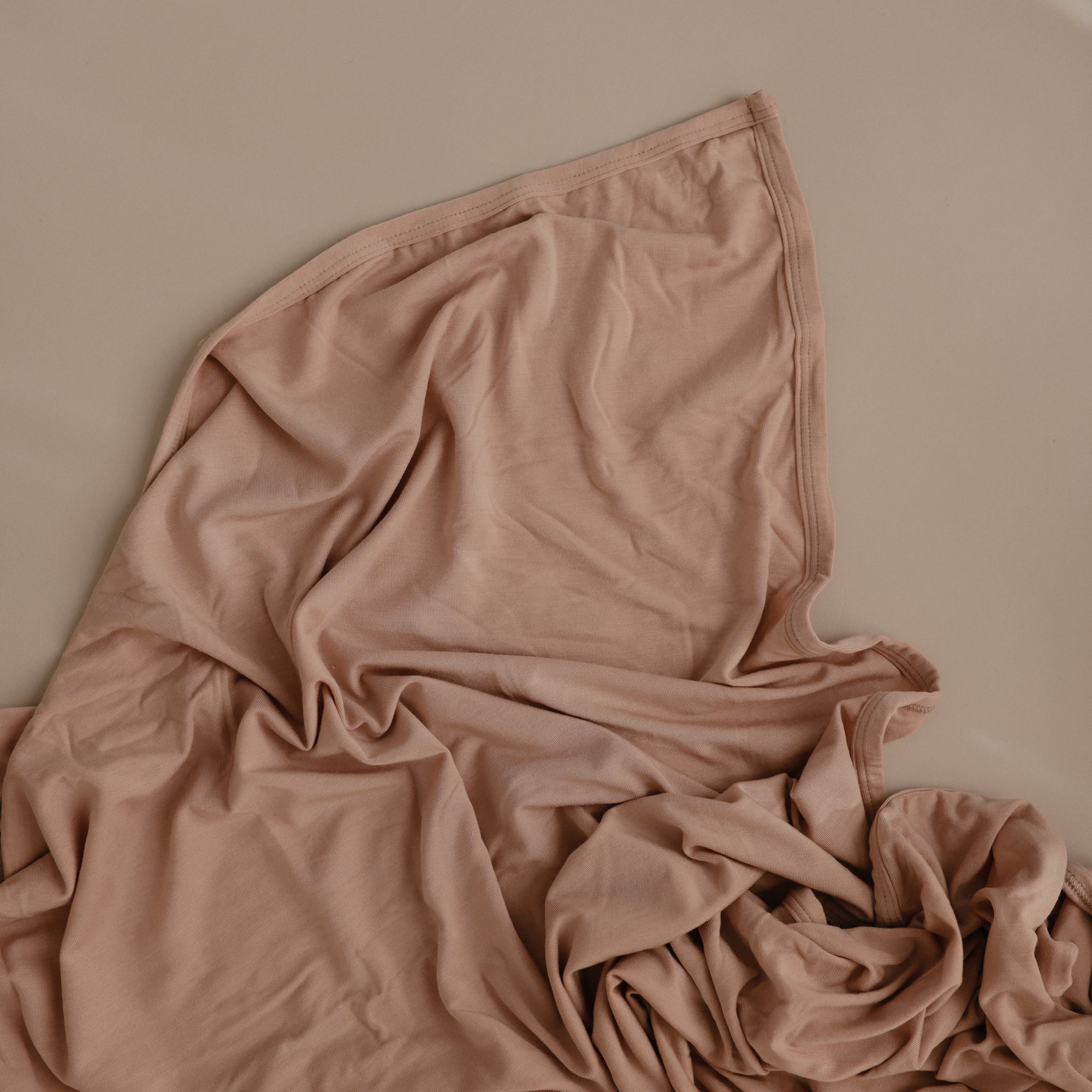 The mushie Stretchy Swaddle, in soft light brown, is gently spread out on a matching beige surface, forming subtle folds and textured lines.