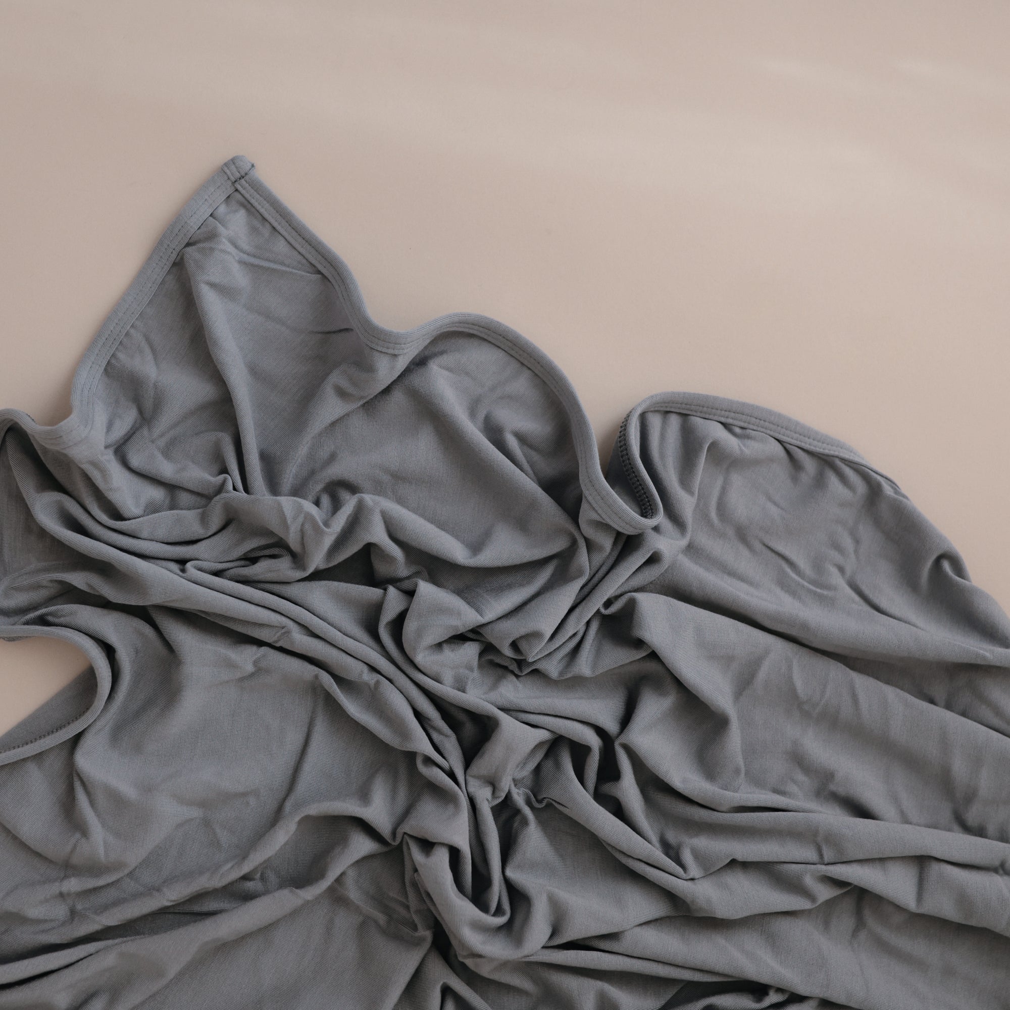 A mushie Stretchy Swaddle in light gray rests crumpled on a smooth beige surface, its breathable fabric forming soft, wavy folds and wrinkles.