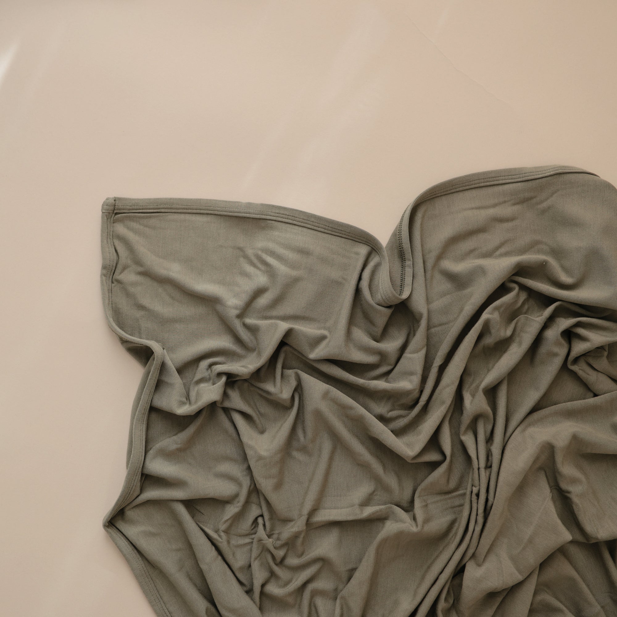 The mushie Stretchy Swaddle, a soft olive-green breathable baby blanket, is loosely spread and wrinkled on a smooth, light beige surface.