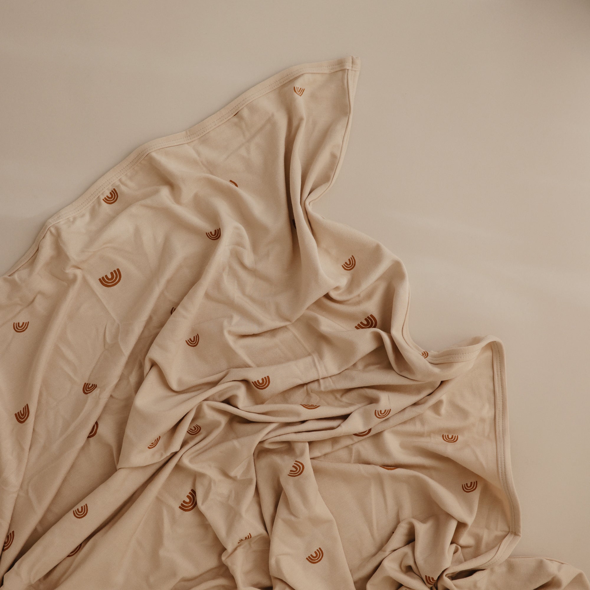The mushie Stretchy Swaddle in light beige, featuring small brown rainbow patterns, is displayed on a matching beige surface with gentle folds highlighting its soft, breathable fabric.