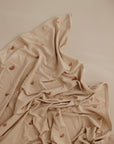 The mushie Stretchy Swaddle in light beige, featuring small brown rainbow patterns, is displayed on a matching beige surface with gentle folds highlighting its soft, breathable fabric.