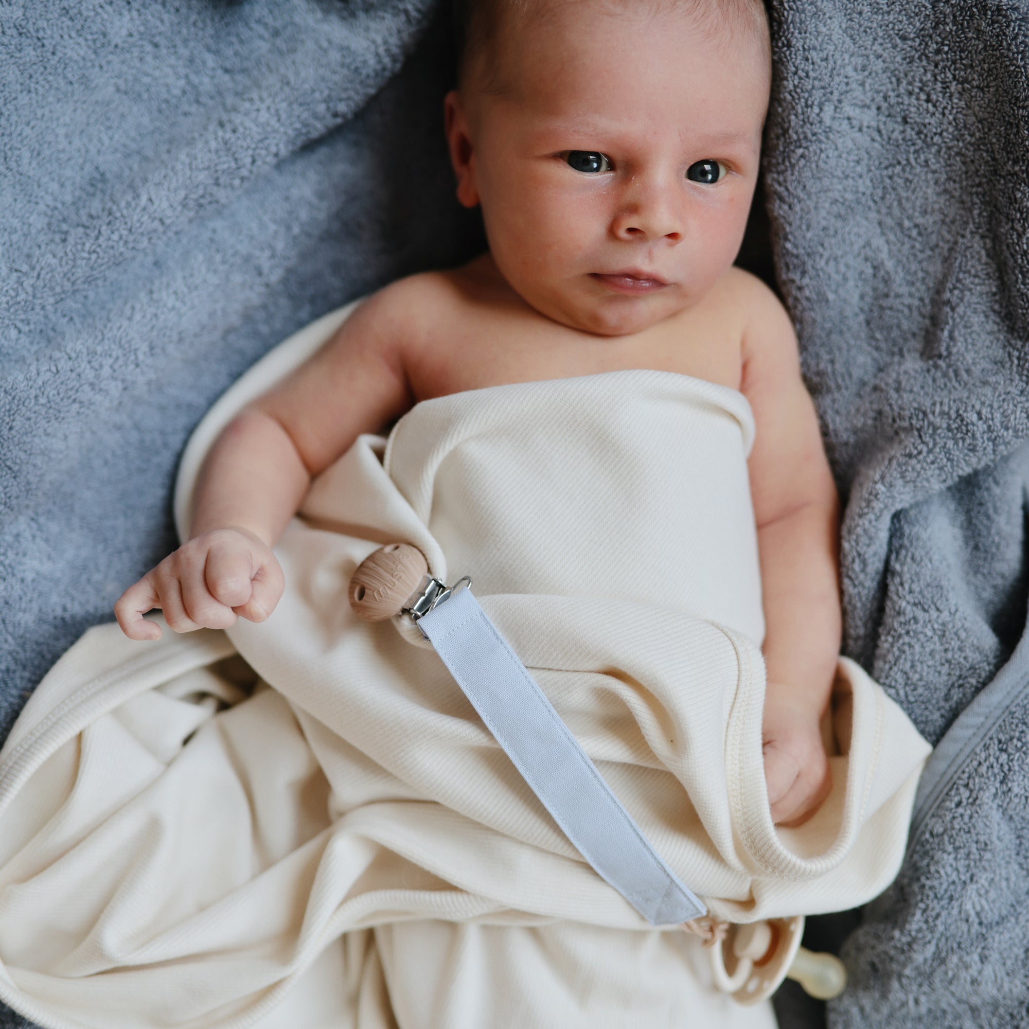 A baby wrapped in a Mushie Ribbed Baby Blanket lies on a soft gray surface, looking up with wide eyes. A pacifier with a wooden clip is attached to the breathable blanket near the baby's hand.