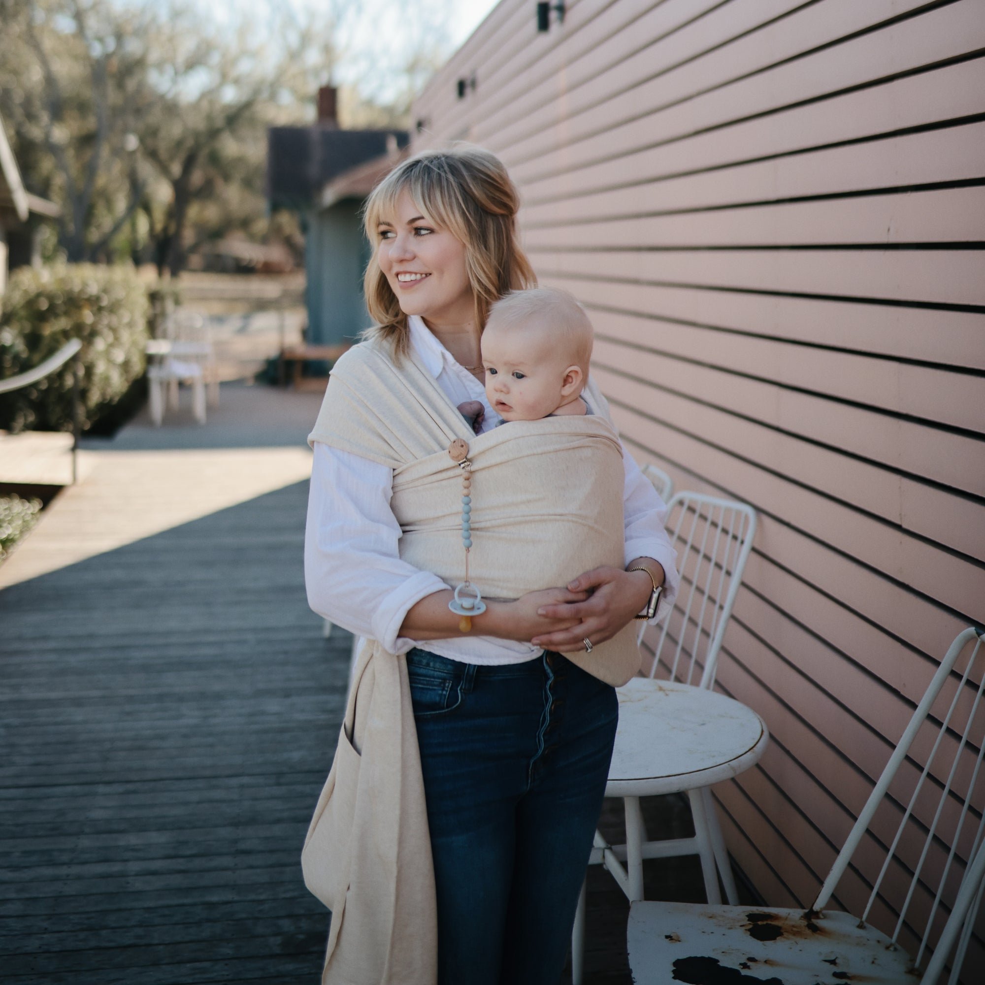 A woman smiles outside by a pink wall, holding a baby in a beige wrap carrier. The baby uses the Mushie FRIGG Rope Natural Rubber Pacifier 2-Pack. Nearby are outdoor chairs, a table, and trees in the background.