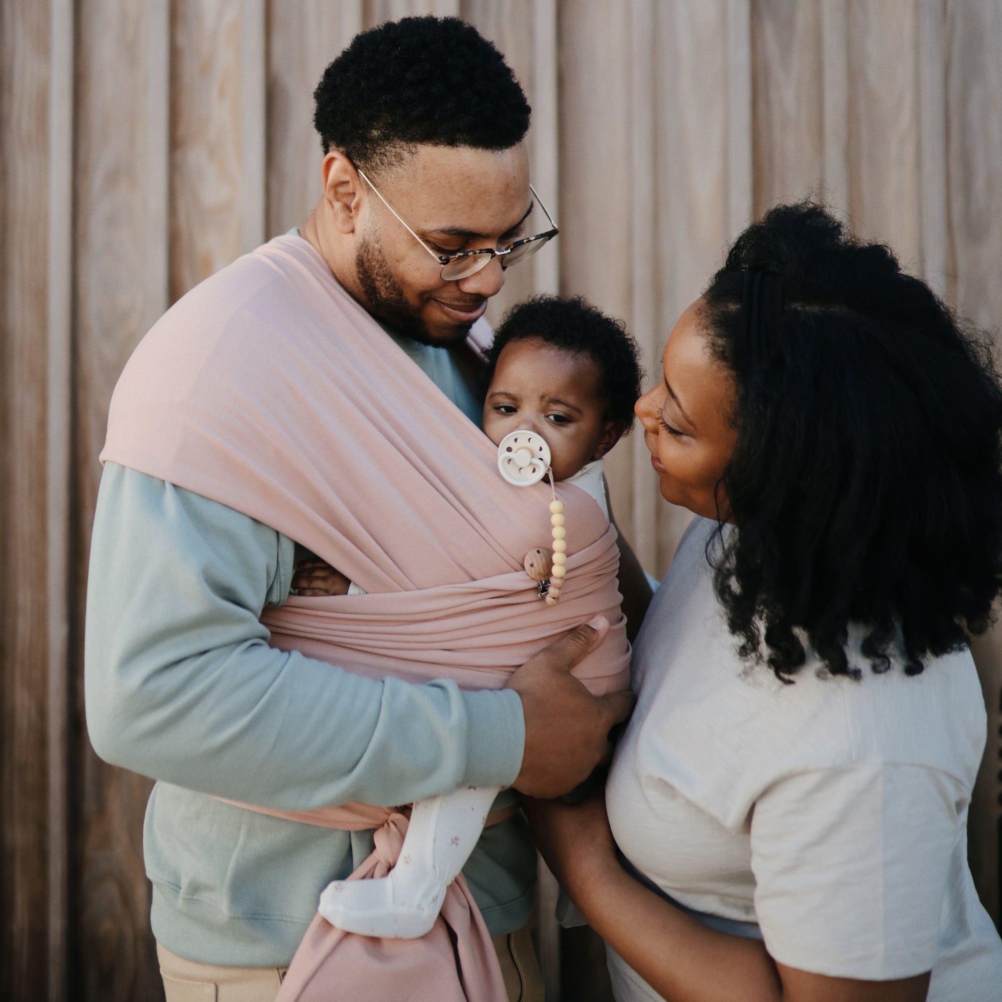 A man holds a baby in a pink wrap carrier, while a woman stands close, smiling. The baby, using the FRIGG Baby's First Pacifier Moonlight Sailing (Cream) 4-Pack by FRIGG, looks at the camera as they stand by a wooden wall.