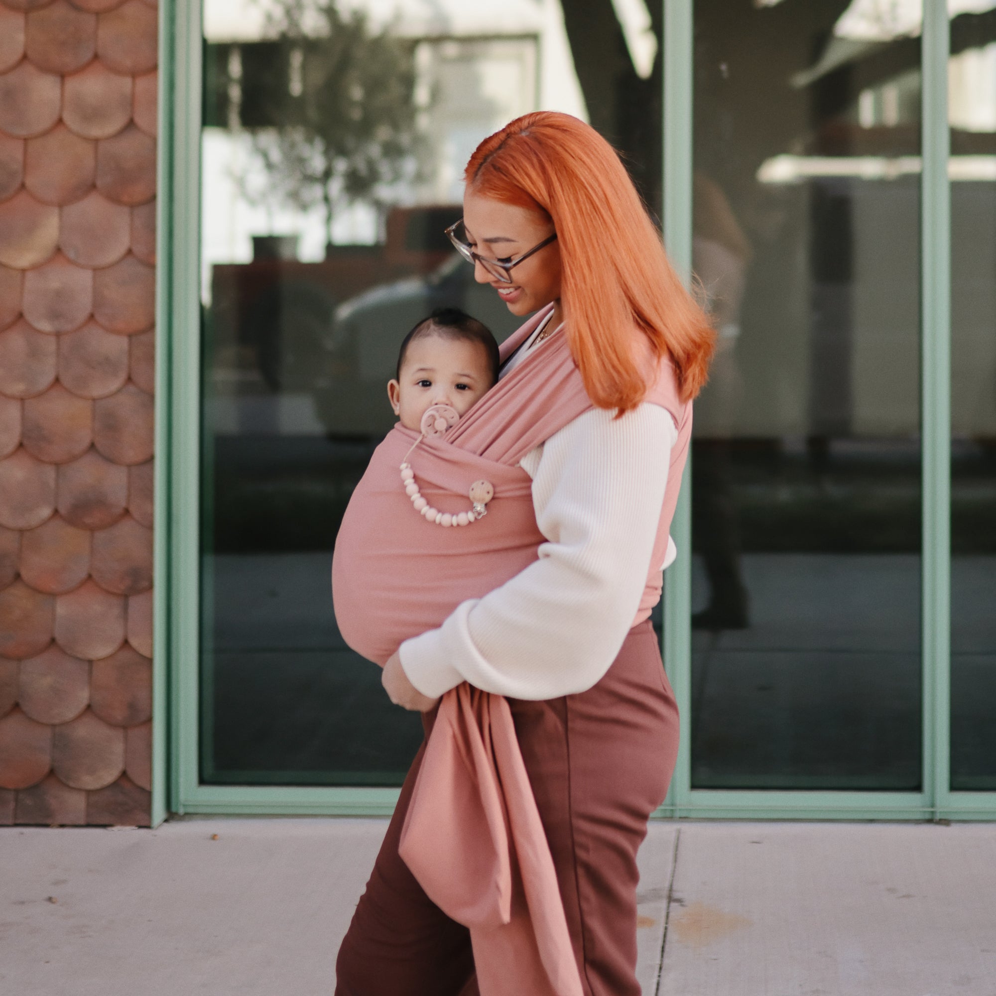 A woman with orange hair and glasses carries a baby in a pink wrap carrier. The baby holds a FRIGG Moon Natural Rubber Pacifier from FRIGG as they stand outside in front of large windows and look at the camera.