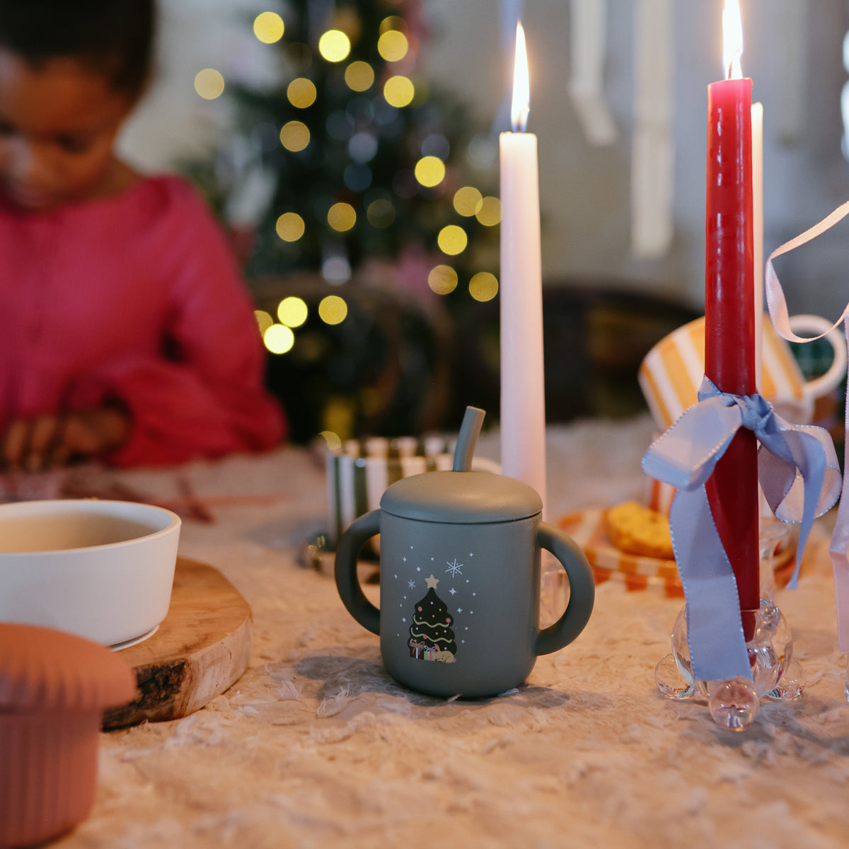 Mushie Silicone Training Cup + Straw with Christmas tree, on table with candles and child behind.