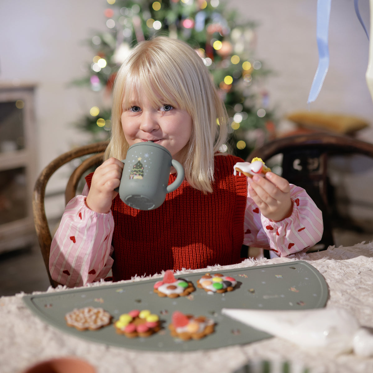 Child with Mushie Silicone Training Cup + Straw enjoys treats by Christmas tree.